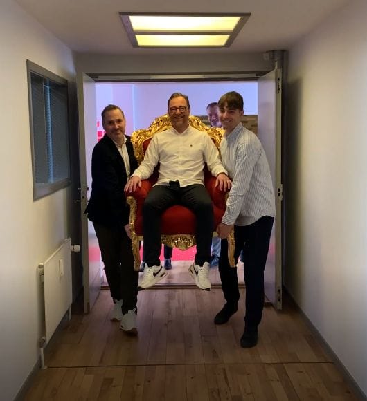 Soren Arnsbo seated on a red and gold throne chair, flanked by two colleagues in an office corridor
